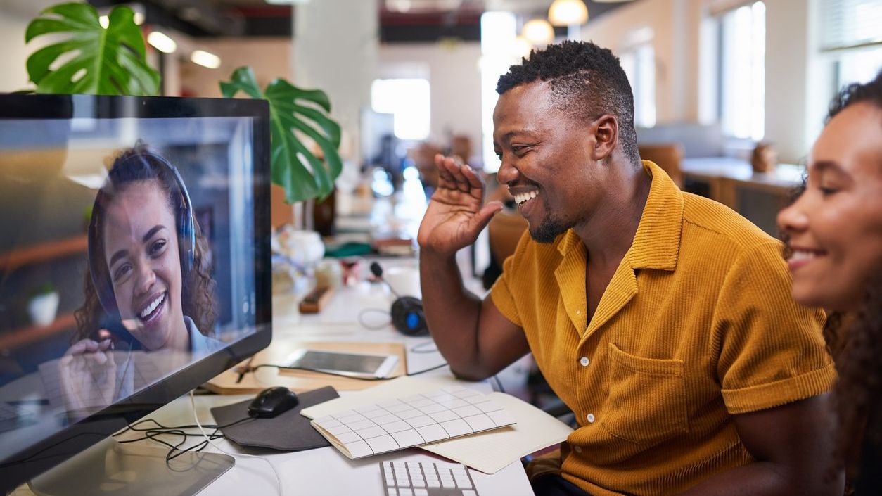 A Black man waves to his colleague on a video call from his office A Black man waves to his colleague on a video call from his office